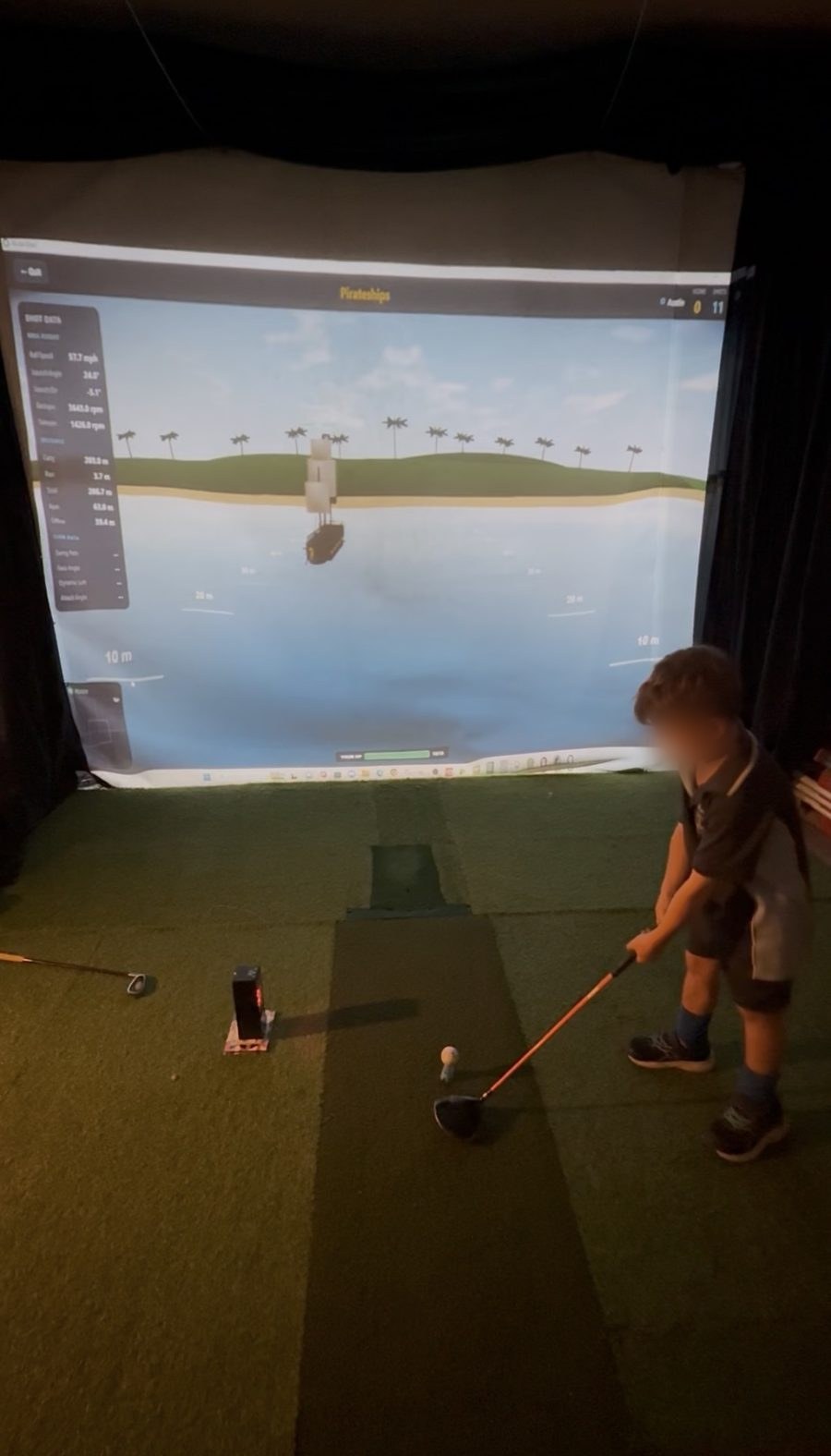 A young boy mid-swing on a golf simulator mat in a garage, playing the Pirateships game on BirdieSmash with live shot data down the left of the screen.